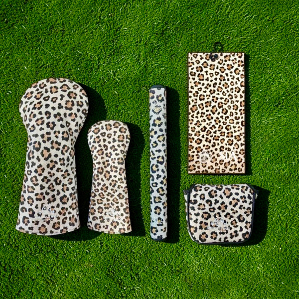 A set of leopard-print golf club covers arranged on bright green grass, including a driver cover, an iron cover, a club shaft cover, a stand, and a bag.