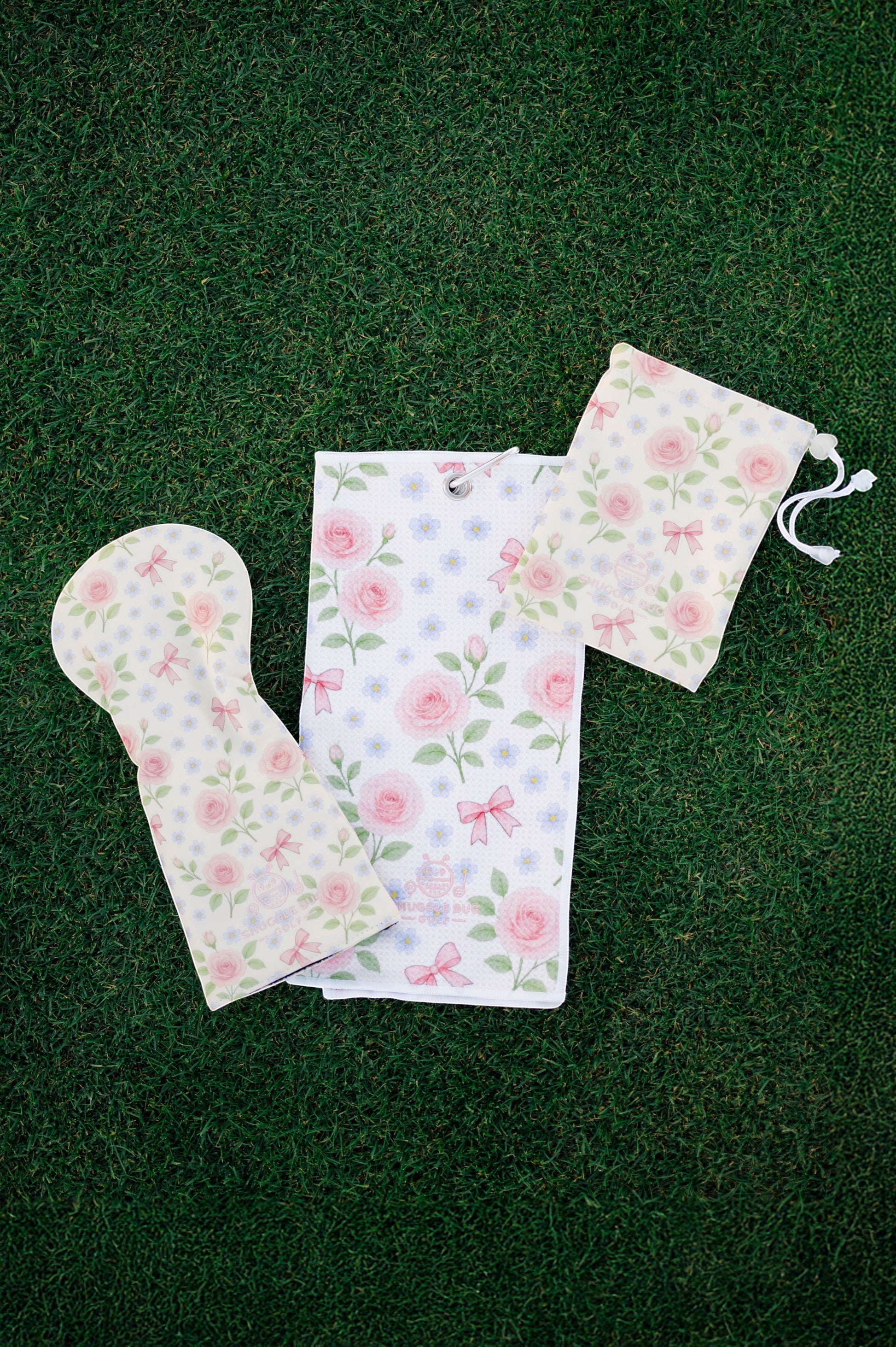 Snuggle Bug Golf Whisper Rose driver head cover, matching towel, and pouch with pink floral and bow print on grass.